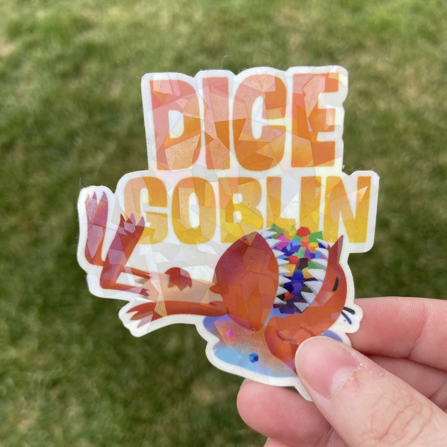 A vinyl sticker of a cartoon goblin laying down with a mouthful of colorful tabletop roleplaying dice. The text reads "DICE GOBLIN" in yellow. The background behind the sticker is dark green.