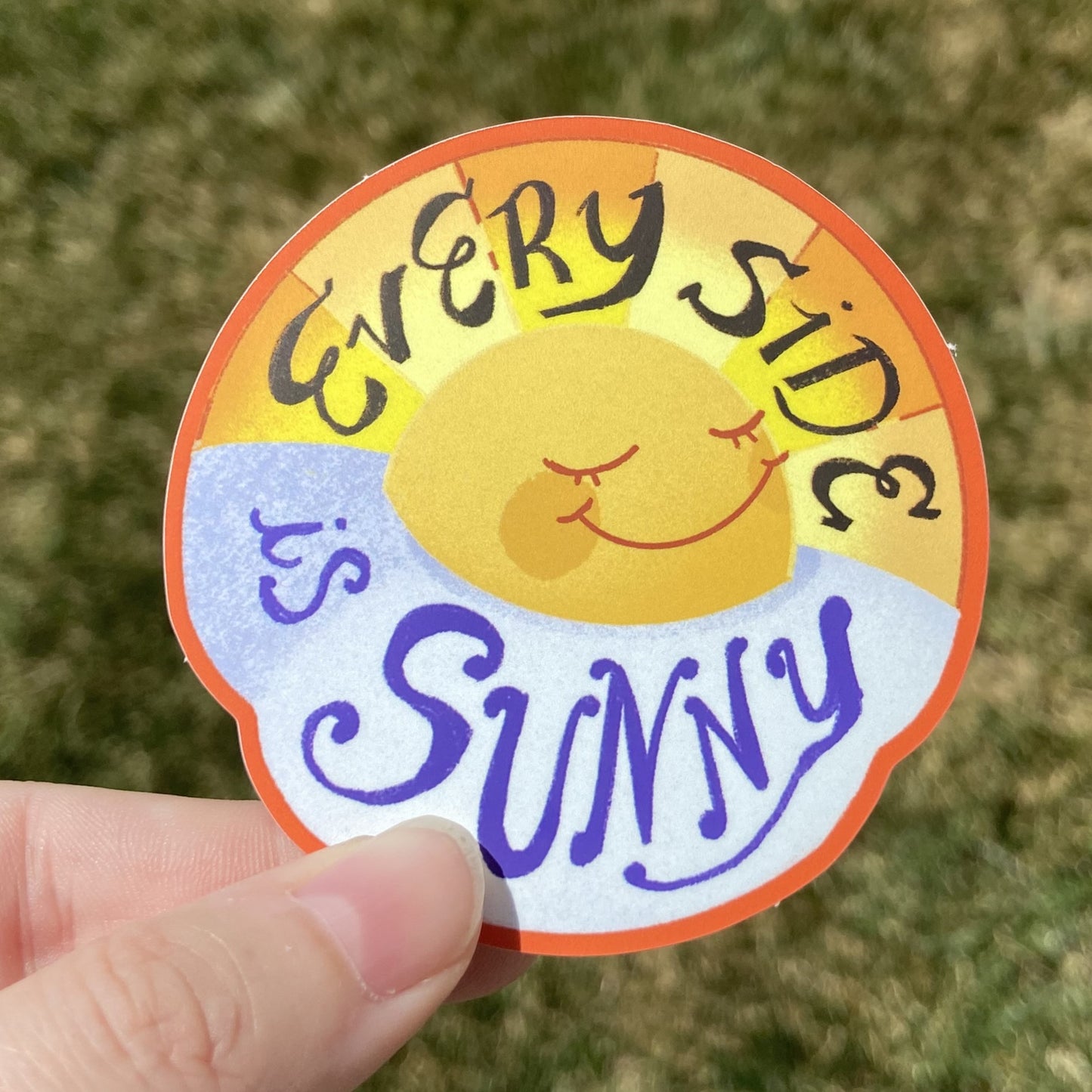 A vinyl sticker featuring an illustration of a cracked egg framed to also resemble a sunrise. There is a sweet smiling face on the yolk. Text on the sticker reads “Every Side’s Sunny” in cursive font.