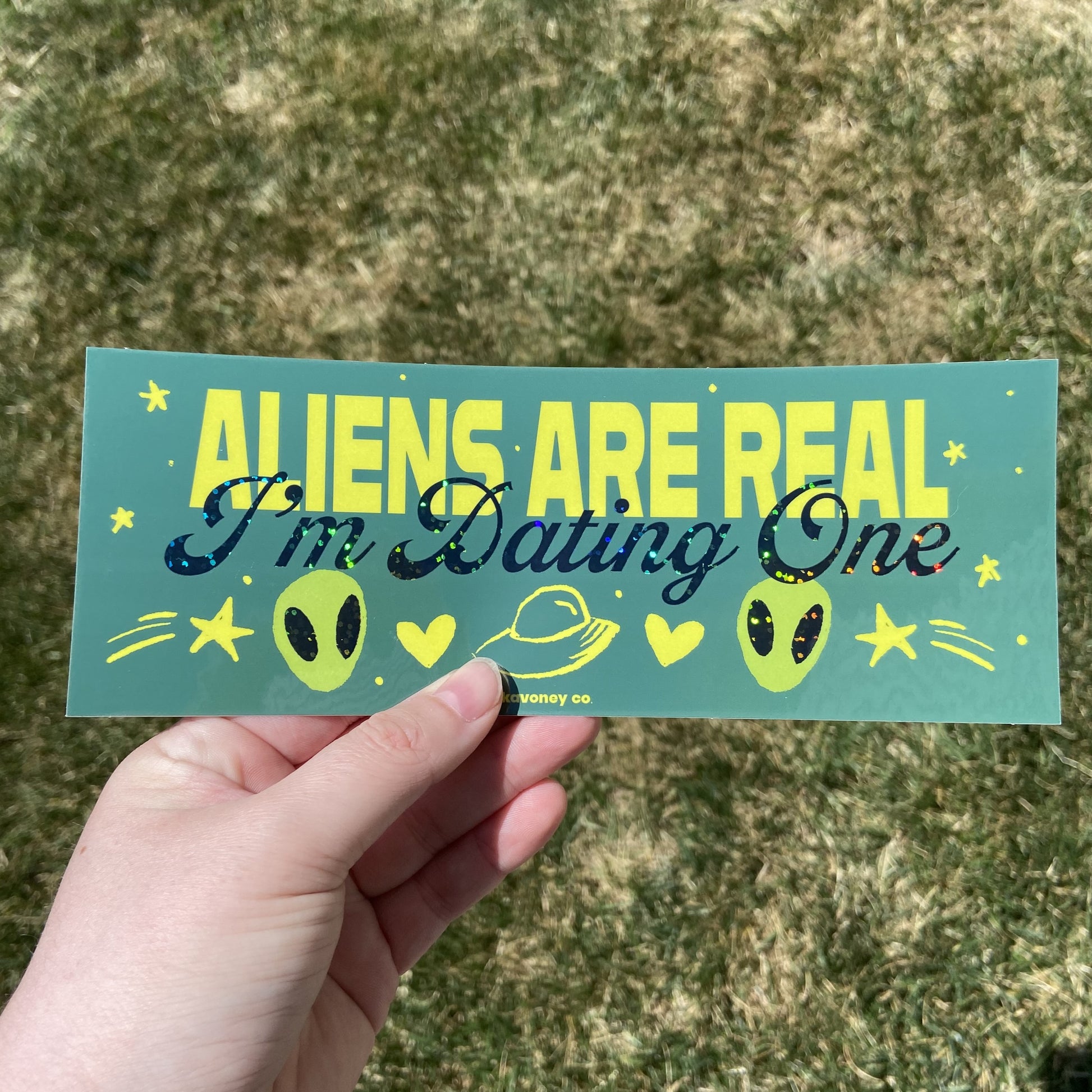 A green rectangular vinyl sticker featuring large yellow text that reads “ALIENS ARE REAL” and then, in smaller dark green glitter cursive text just below, “I’m Dating One.” Below the text is a graphic of two small green alien creatures, a space ship, two yellow hearts, and two yellow shooting stars.