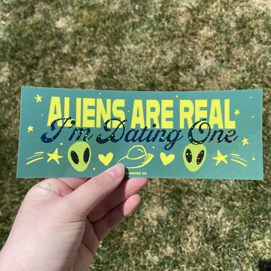 A green rectangular vinyl sticker featuring large yellow text that reads “ALIENS ARE REAL” and then, in smaller dark green glitter cursive text just below, “I’m Dating One.” Below the text is a graphic of two small green alien creatures, a space ship, two yellow hearts, and two yellow shooting stars.