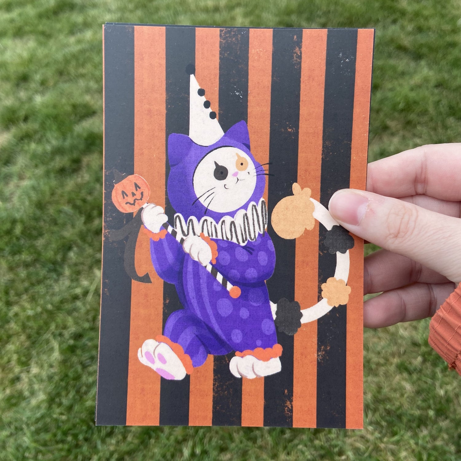 A postcard oriented vertically, featuring a spot illustration of a calico cat dressed as a Halloween-themed clown in a purple costume. The background behind the cat is black and orange vertical stripes. The background behind the postcard is dark green.