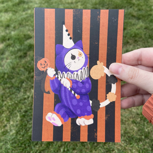 A postcard oriented vertically, featuring a spot illustration of a calico cat dressed as a Halloween-themed clown in a purple costume. The background behind the cat is black and orange vertical stripes. The background behind the postcard is dark green.