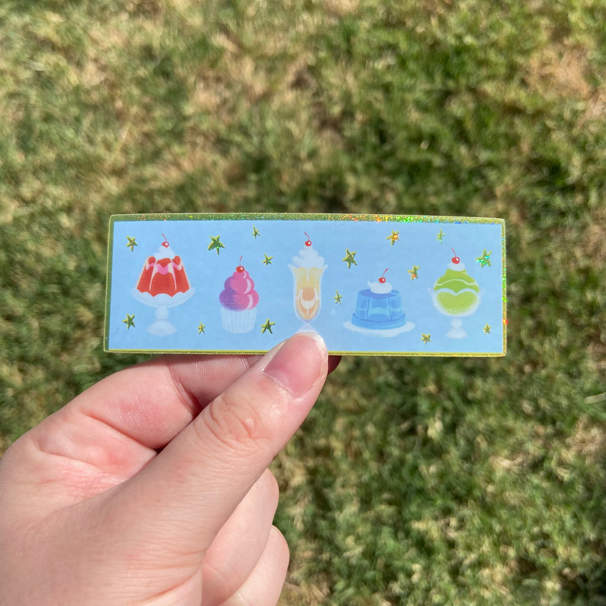 A rectangular vinyl sticker featuring illustrations of five colorful dessert items lined up in a row. The background behind the desserts is baby blue with glittering yellow stars and a matching yellow glitter border.
