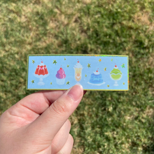 A rectangular vinyl sticker featuring illustrations of five colorful dessert items lined up in a row. The background behind the desserts is baby blue with glittering yellow stars and a matching yellow glitter border.