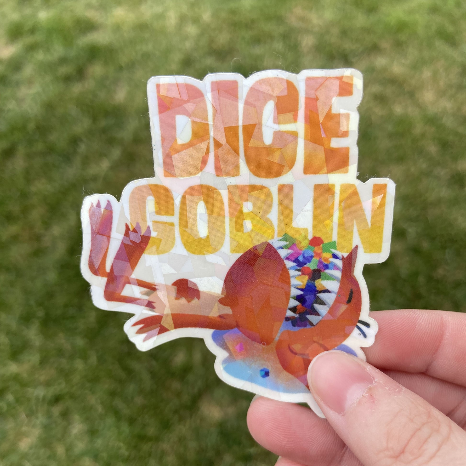 A vinyl sticker of a cartoon goblin laying down with a mouthful of colorful tabletop roleplaying dice. The text reads "DICE GOBLIN" in yellow. The background behind the sticker is dark green.