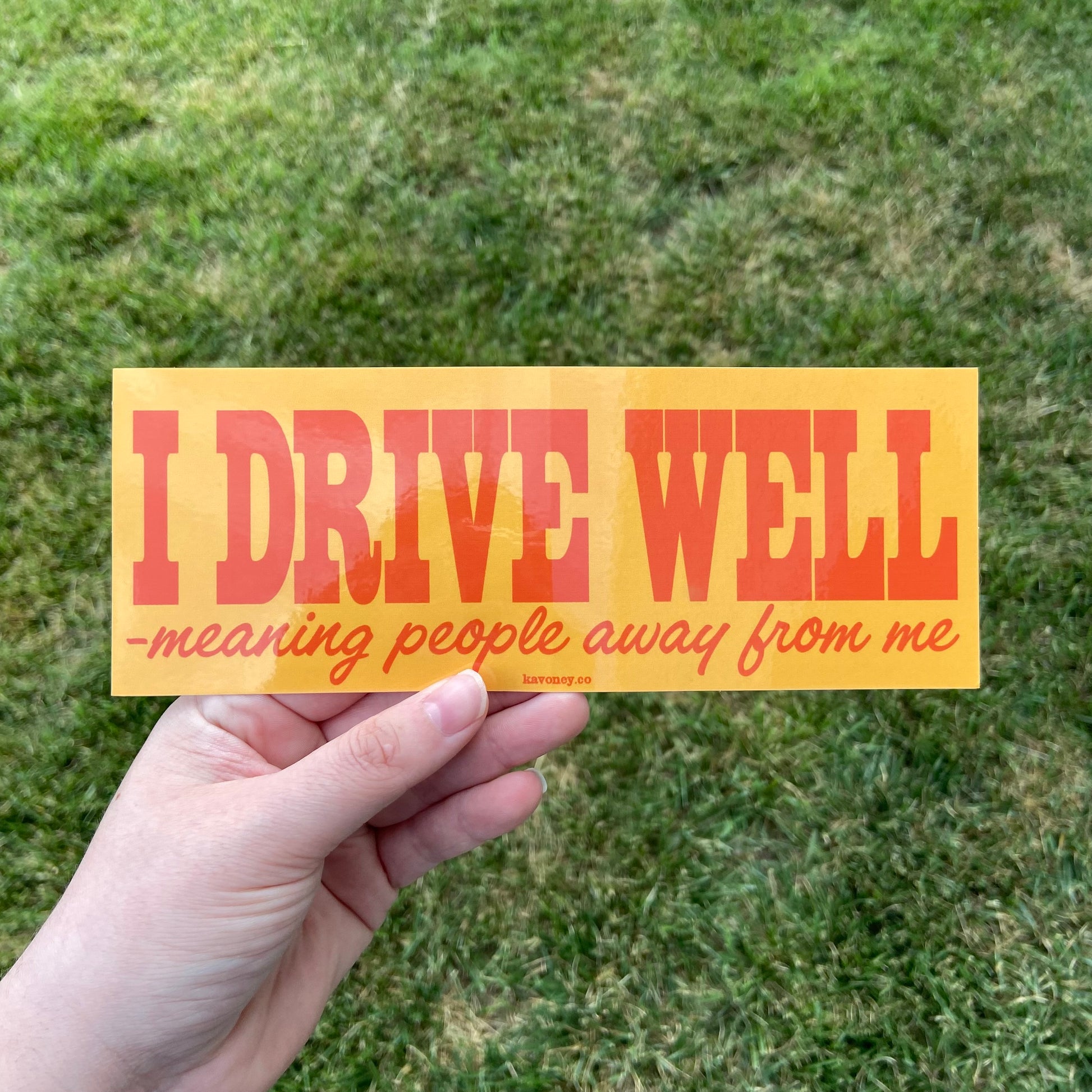 A warm yellow rectangular vinyl sticker featuring large text that reads “I DRIVE WELL” in orange font. Below it, in smaller font, the rest of the text reads “-meaning people away from me” as a continuation of the sentence.