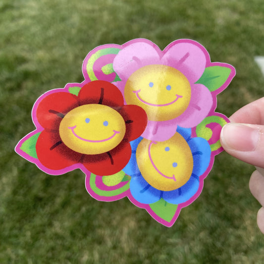 A vinyl sticker of three smiling flowers, one pink, one red, and one blue, with vine and leaf details. The background behind the sticker is dark green.