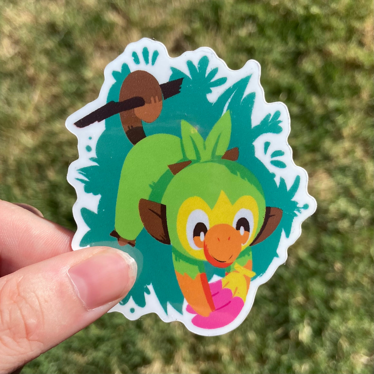 A vinyl sticker featuring an illustration of a cute green grass monkey monster! It is hanging by its tail from a branch and grabbing a pink banana-shaped fruit.
