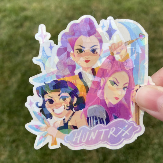 A vinyl sticker featuring three characters from K-Pop Demon Hunters, Rumi, Mira, and Zoey, holding their weapons. Text at the bottom of the sticker reads "Huntrix" in the logo font. 