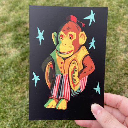 A postcard oriented vertically. The postcard features a spot illustration of a vintage cymbal clapping monkey toy, colored in shades of red and yellow and surrounded by teal stars in front of a black background. The background behind the postcard is dark green.
