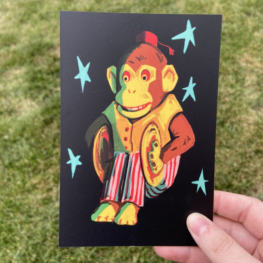 A postcard oriented vertically. The postcard features a spot illustration of a vintage cymbal clapping monkey toy, colored in shades of red and yellow and surrounded by teal stars in front of a black background. The background behind the postcard is dark green.
