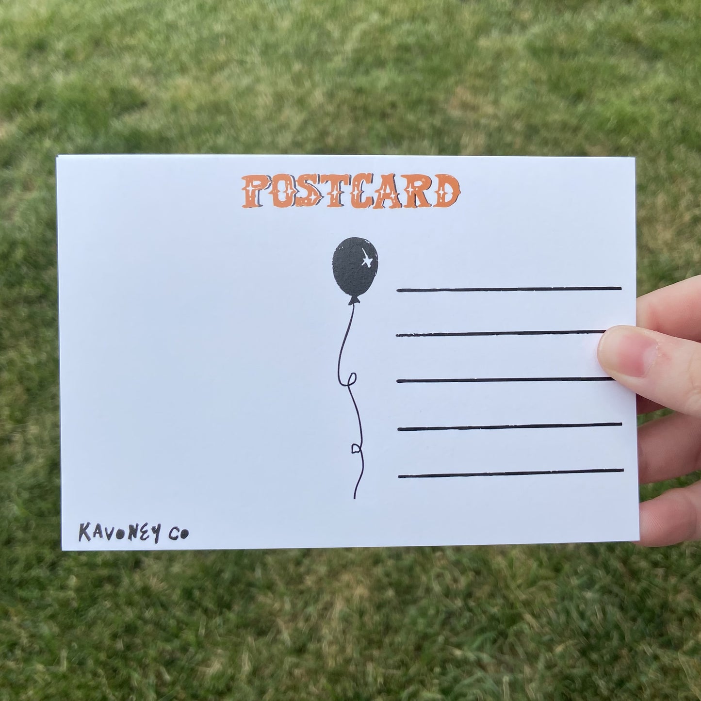 The back of a postcard oriented horizontally. The top text reads"Postcard" in an embellished font. There are lines for writing, and a balloon graphic in the center. The bottom left corner says "Kavoney Co." The background behind the postcard is dark green.