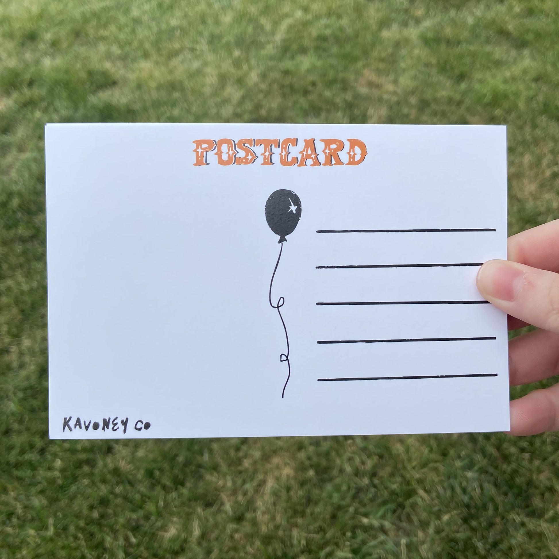 The back of a postcard oriented horizontally. The top text reads"Postcard" in an embellished font. There are lines for writing, and a balloon graphic in the center. The bottom left corner says "Kavoney Co." The background behind the postcard is dark green.