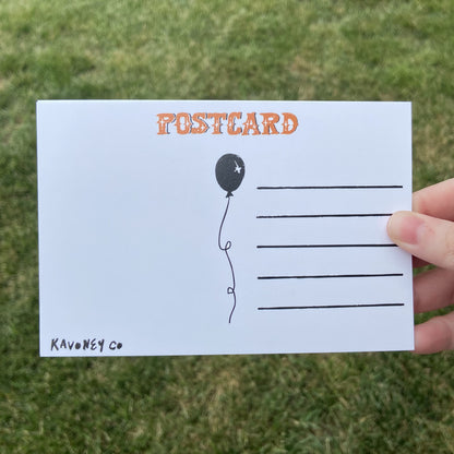 The back of a postcard oriented horizontally. The top text reads"Postcard" in an embellished font. There are lines for writing, and a balloon graphic in the center. The bottom left corner says "Kavoney Co." The background behind the postcard is dark green.