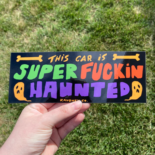A black rectangular vinyl sticker featuring bold handlettered text that reads “This Car Is Super Fuckin’ Haunted” in yellow, orange, green, and purple. There are two illustrated bones and two illustrated ghosts embellishing the text at the top and bottom.