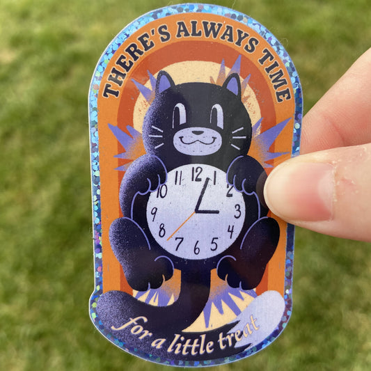 A vinyl sticker of a black and white cat clock over a colorful oval-shaped background in shades of orange. The text on the sticker reads "There's Always Time for a little treat". The border of the sticker is glittery blue. The background behind the sticker is dark green.